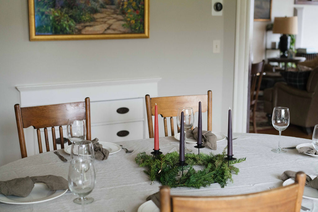 A dining table set for a meal with a white tablecloth, wooden chairs, wine glasses, gray napkins, and an evergreen Advent wreath with Schneider Goods beeswax taper candles.
