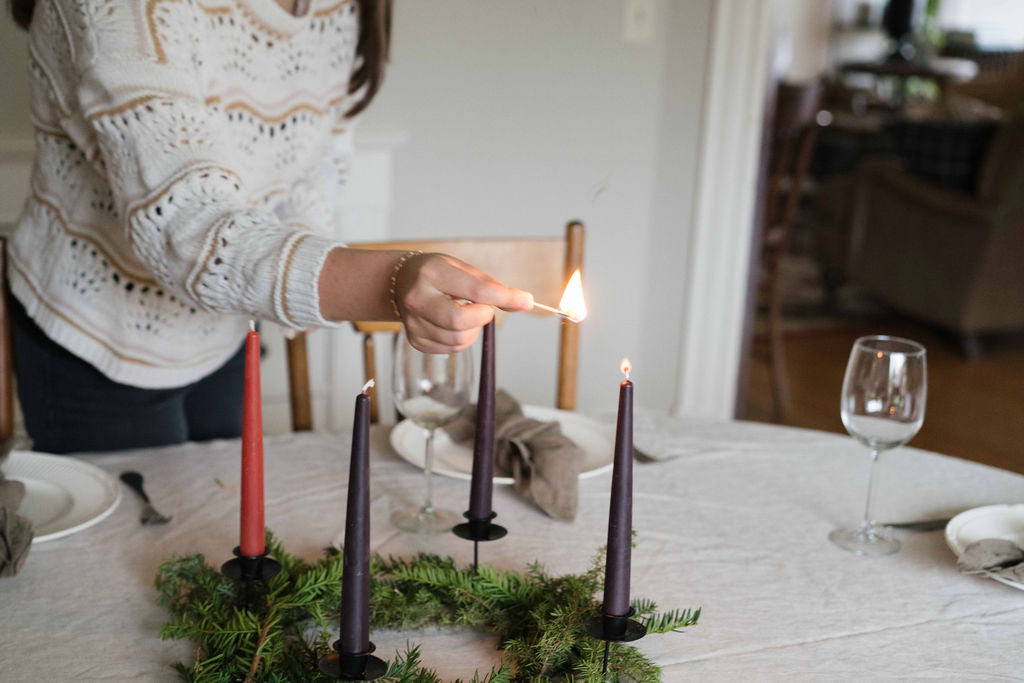 Woman in a cream knit sweater lights a dark purple Advent taper candle on a dining table.