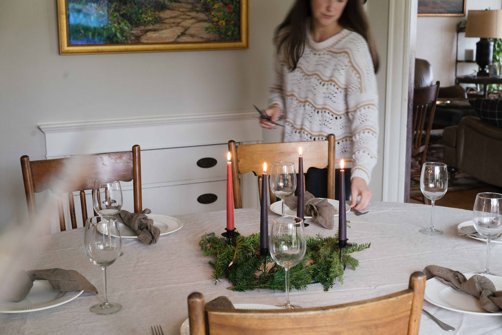 A woman in a cream knit sweater stands beside a dining table set with white plates, wine glasses, folded gray napkins, and a green Advent wreath with beeswax candles.