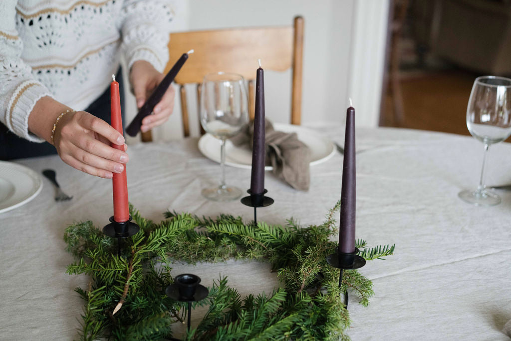 A person's hands setting a pink taper candle in a black metal holder, set within a green pine wreath centerpiece on a white tablecloth. Other dark purple taper candles and wine glasses are visible on a dining table.