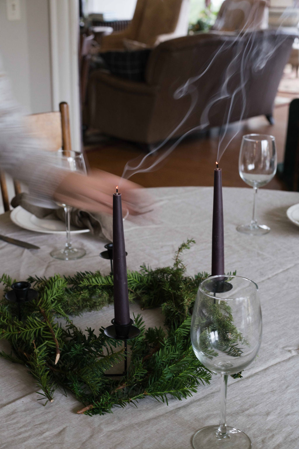A dining table set with a green Advent wreath holding Schneider Goods beeswax Advent candles, with smoke rising from the blown-out candles. Clear wine glasses and plates are arranged around the table in a home dining room setting.