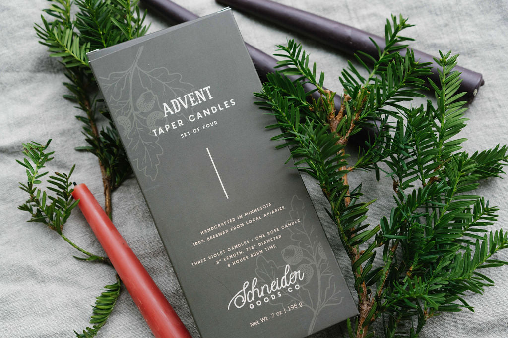 Schneider Goods Advent Taper Candles set of four, featuring a dark green box surrounded by green pine branches and candles in pink and dark purple on a gray fabric background.