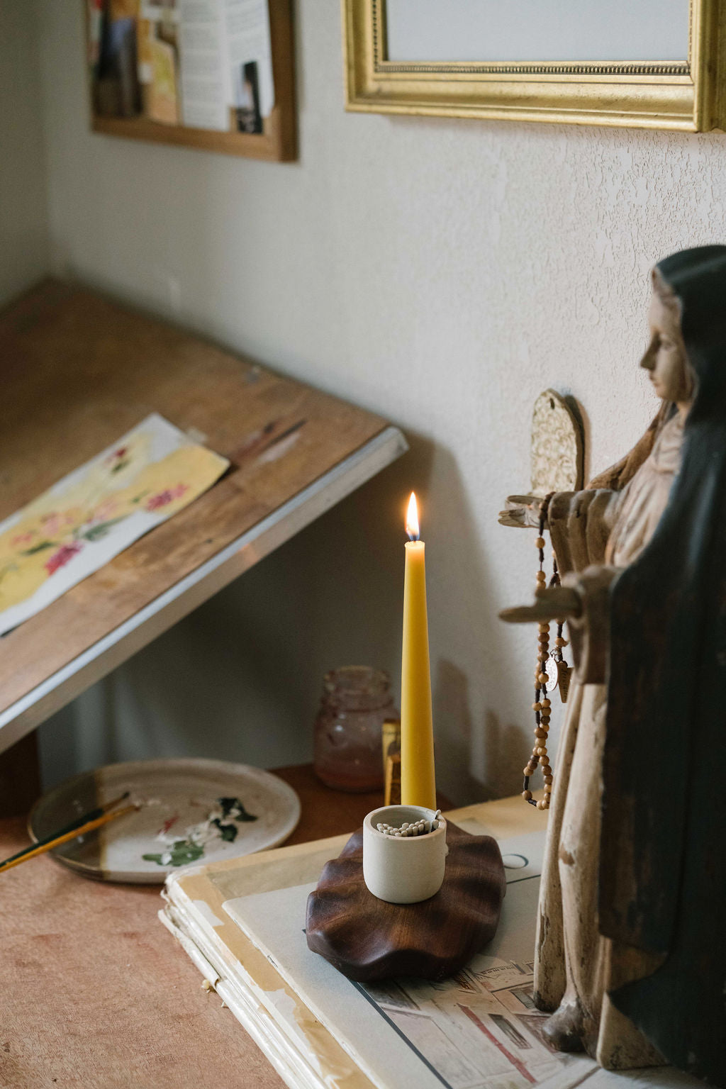 A lit yellow taper candle in a small holder sits on a wooden surface next to a statue of Mary and rosary in a softly lit room.