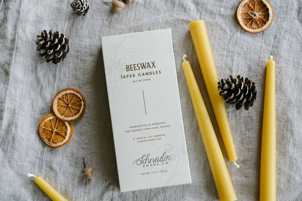 Box of four beeswax taper candles on a beige background with pinecones and orange slices.