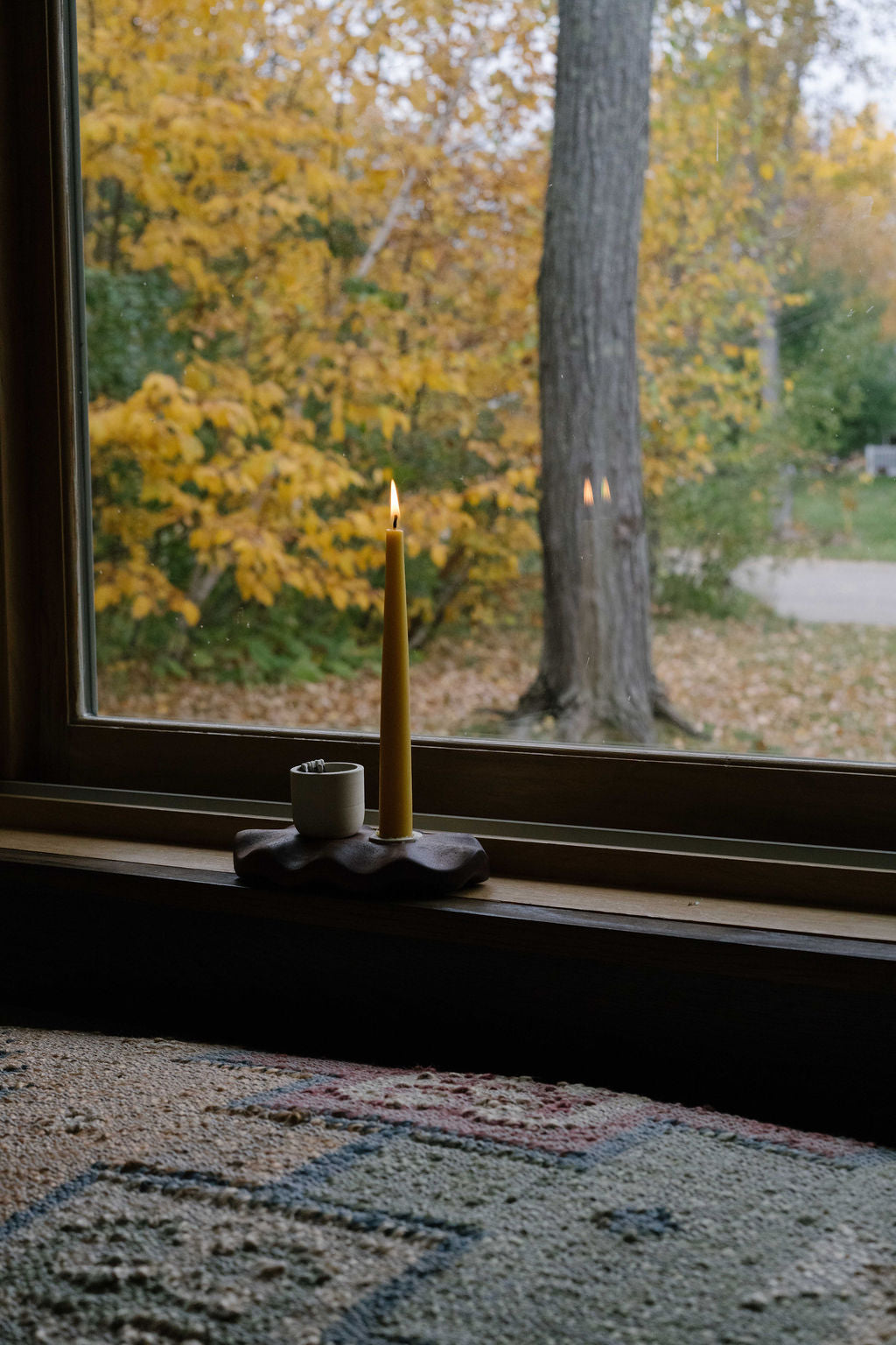 A single beeswax taper candle lit in a windowsill. The window behind shows yellow fall leaves.