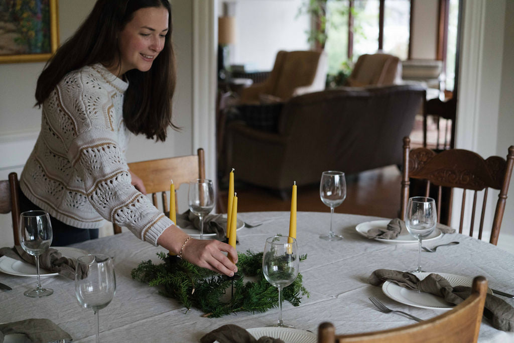 Woman putting four beeswax candles into an evergreen wreath in the middle of a dining table.