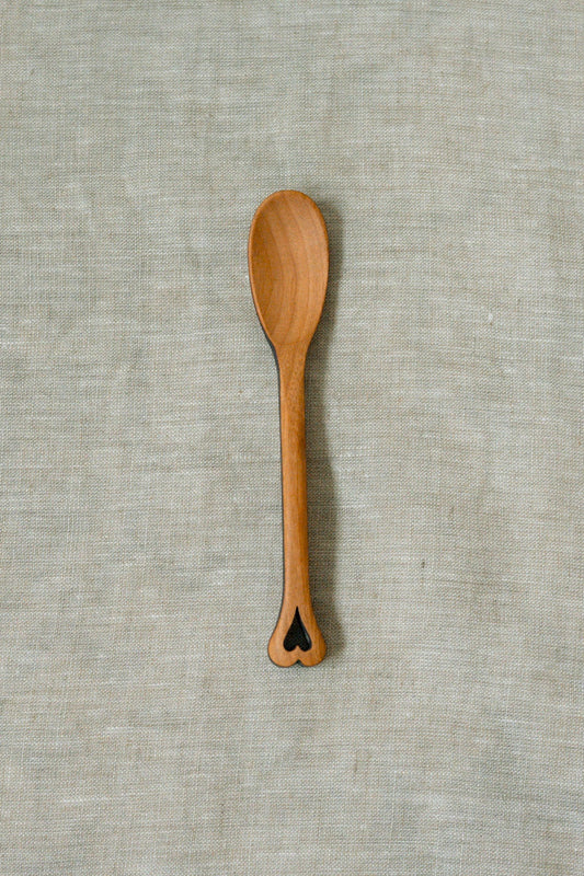 A small wooden spoon with a heart-shaped cutout at the end of the handle, placed vertically on a textured light beige linen fabric background.