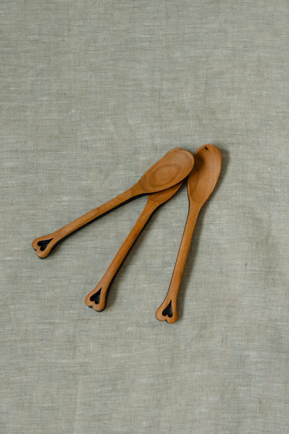 Three small wooden spoons with decorative cutout handles, arranged diagonally on a textured light gray linen fabric background.