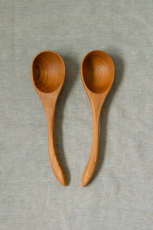 Two light brown wooden ladles with long handles, displayed side-by-side on a textured light gray linen background.
