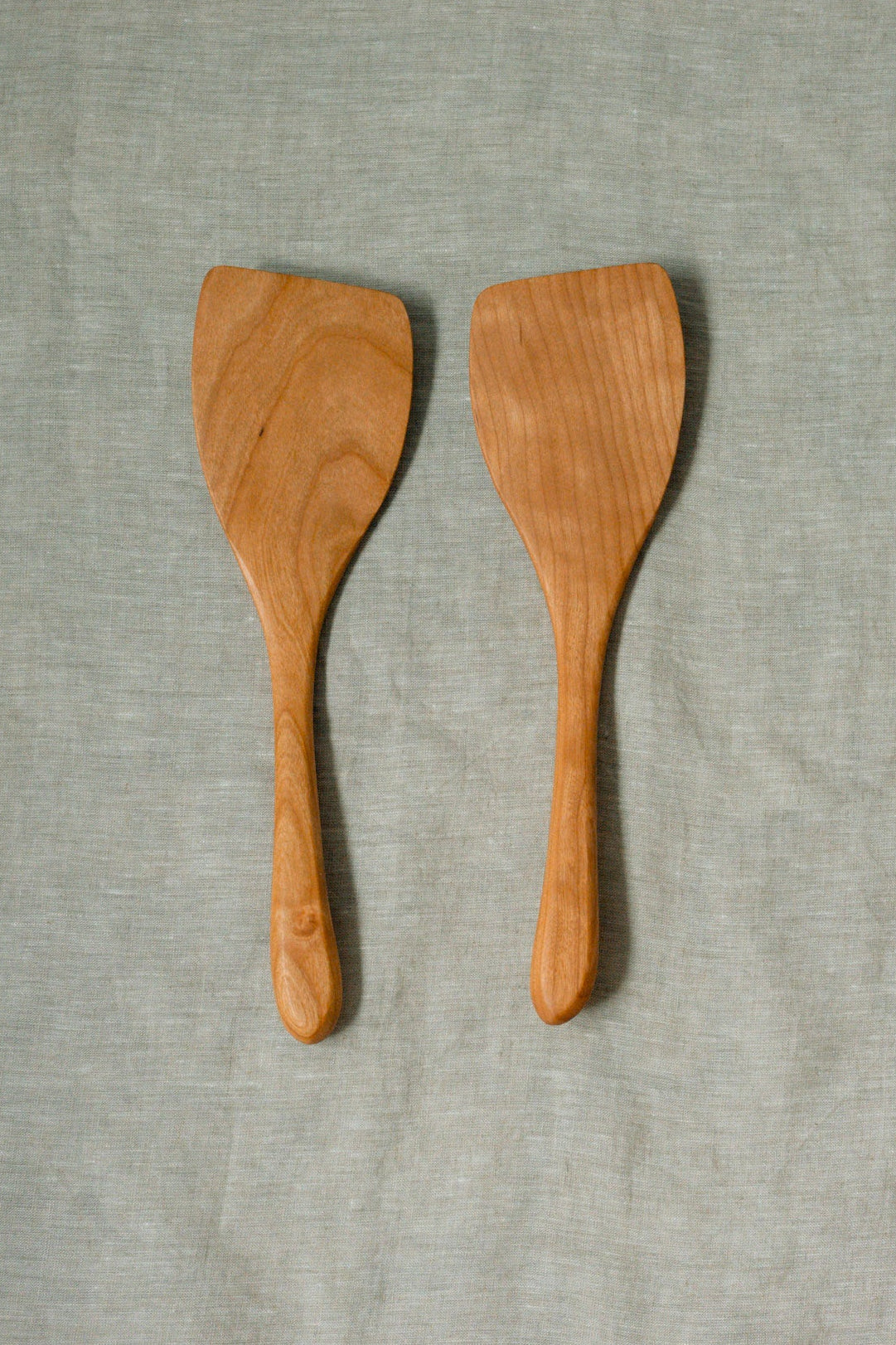 Two light brown wooden spatulas with visible wood grain, displayed side-by-side on a textured light gray fabric background.