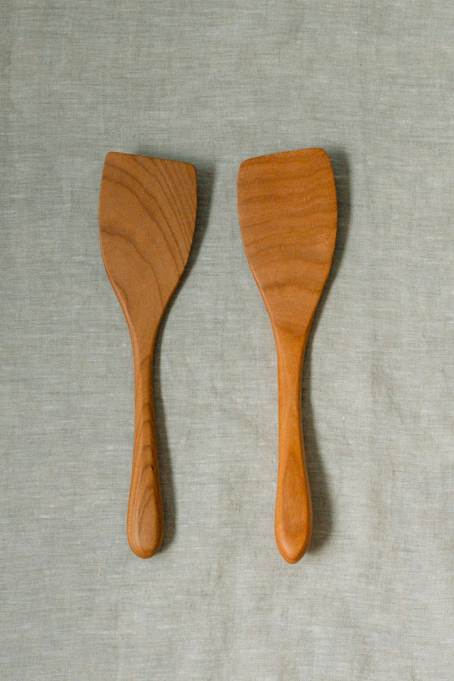 Two light brown wooden spatulas with visible wood grain, displayed side-by-side on a textured light gray linen fabric background.