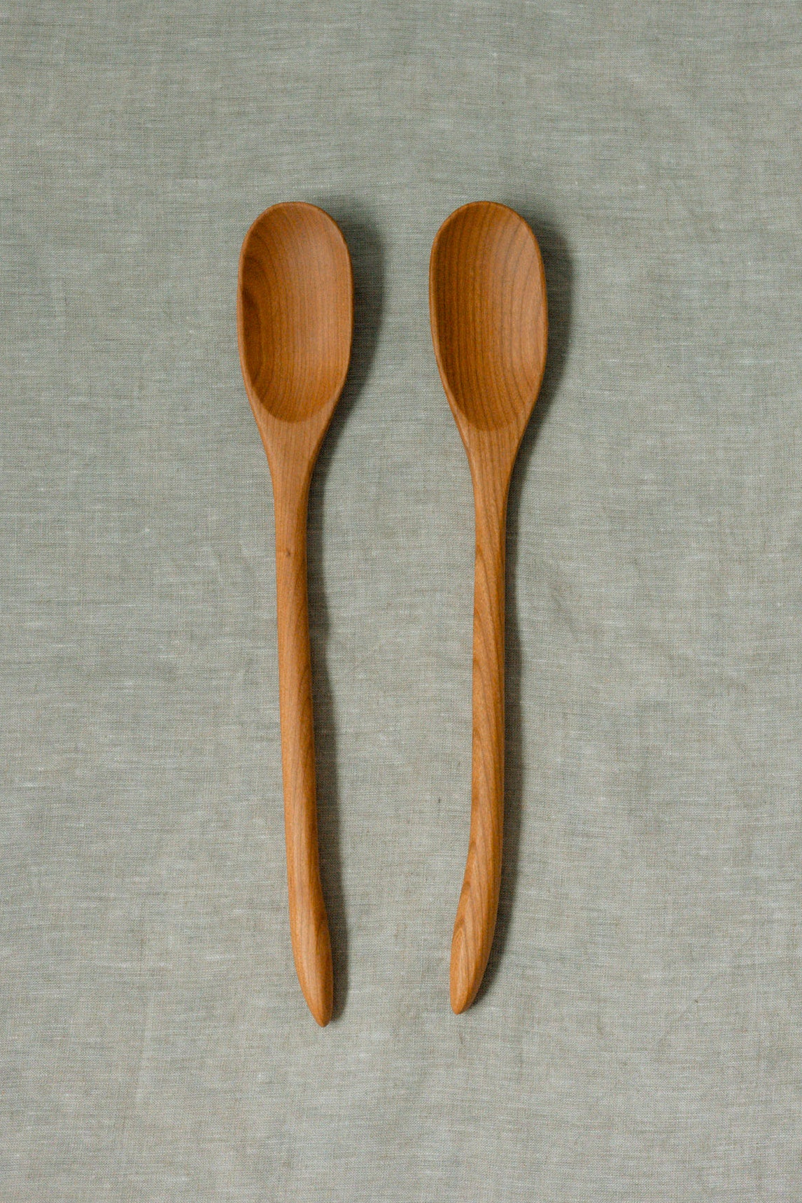 Two light brown wooden spoons with visible wood grain, arranged vertically side-by-side on a textured light gray linen fabric background.