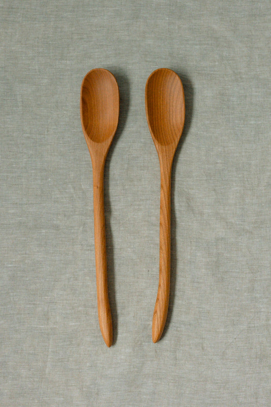 Two light brown wooden spoons with visible wood grain, arranged vertically side-by-side on a textured light gray linen fabric background.