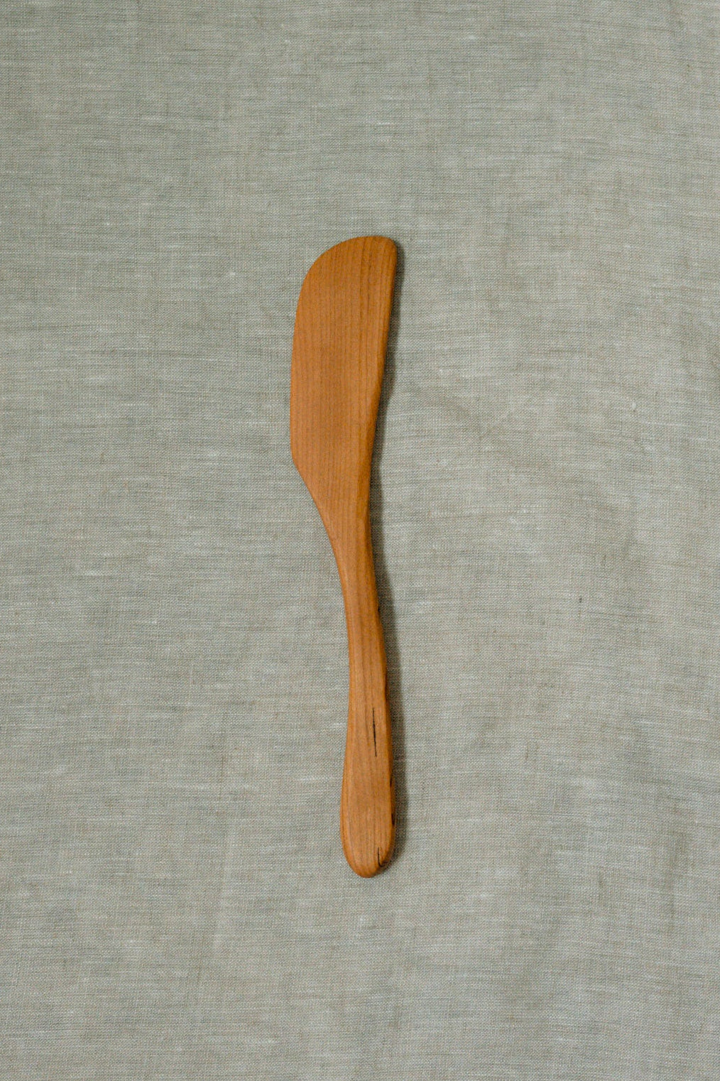 A small wooden spatula or spreader with a smooth, light brown finish, placed vertically on a textured light gray linen fabric background.