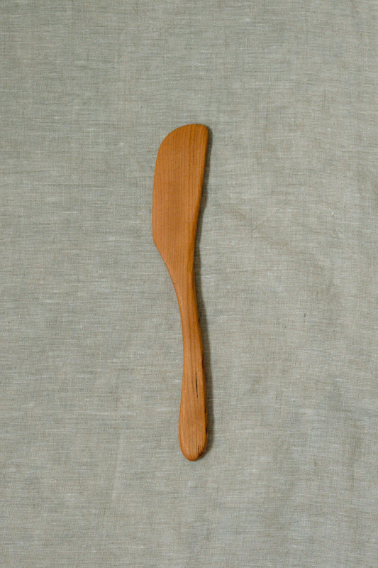 A small wooden spatula or spreader with a smooth, light brown finish, placed vertically on a textured light gray linen fabric background.