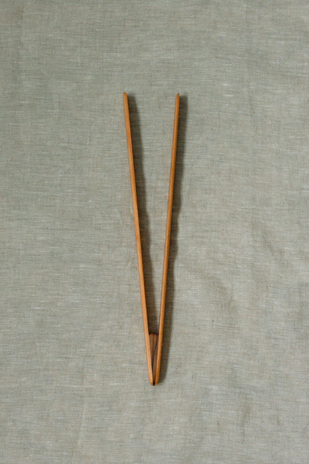 A pair of light brown wooden tongs, displayed on a textured light gray linen background.