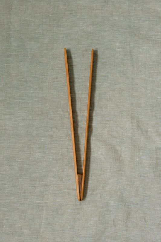 A pair of light brown wooden tongs, displayed on a textured light gray linen background.