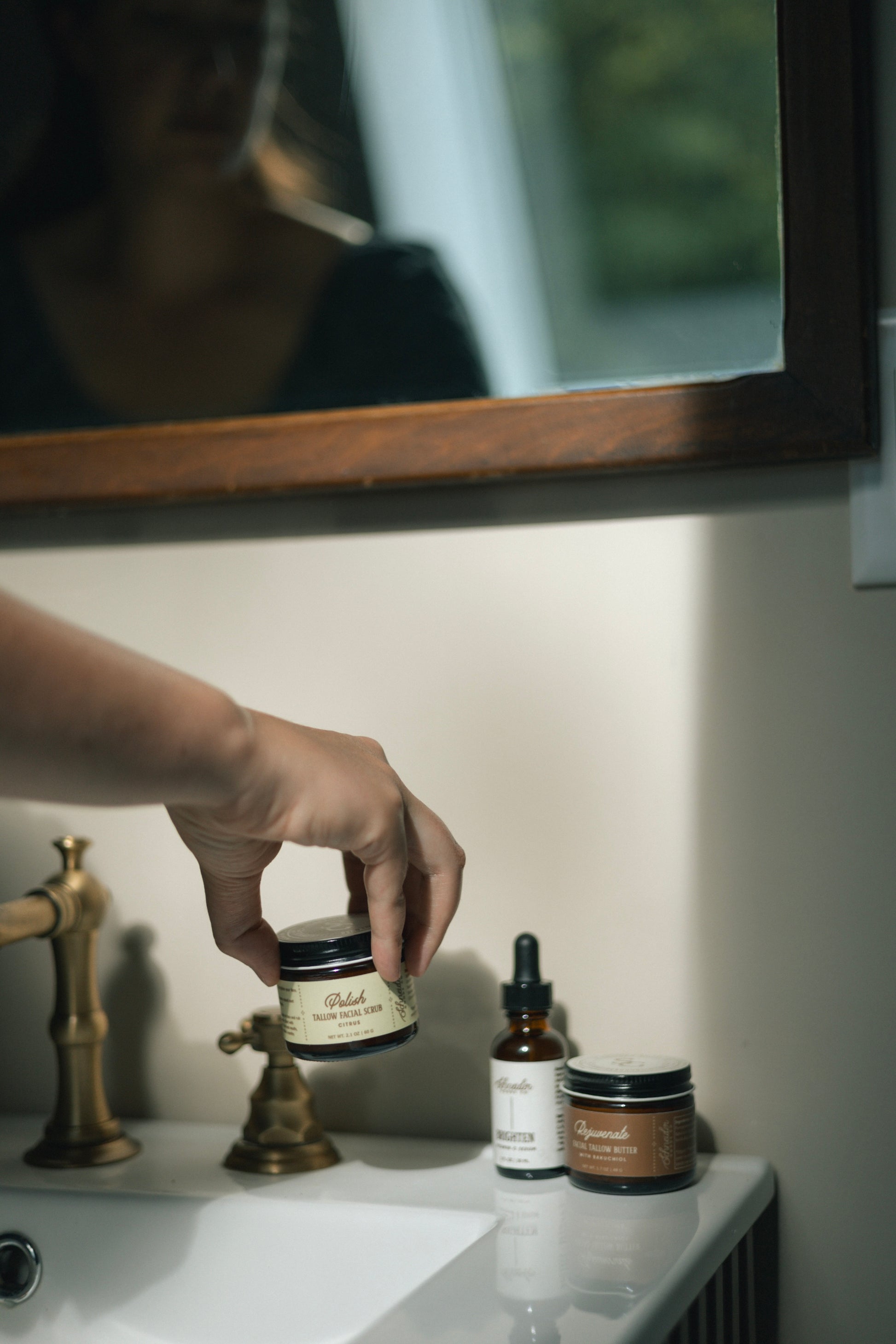 A hand places Polish Facial Scrub on a white counter, alongside Brighten Vitamin C Serum and Rejuvenate Facial Tallow.