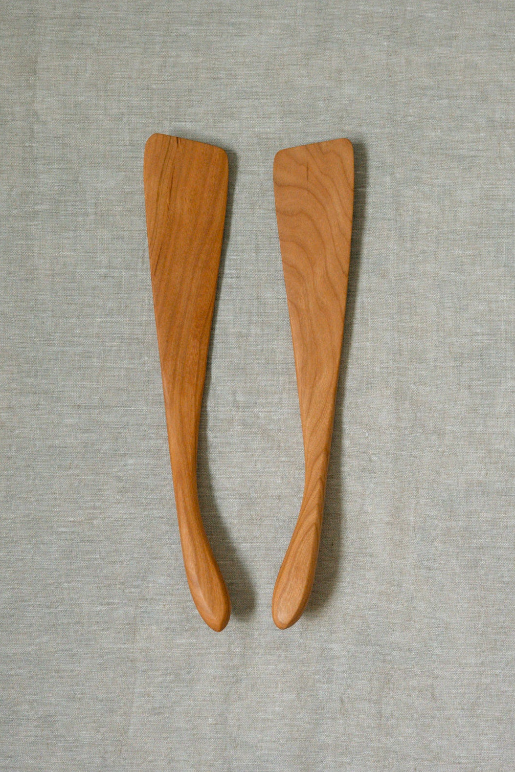 Two cherry wooden french spatulas with visible wood grain, placed side-by-side on a textured light gray fabric background.