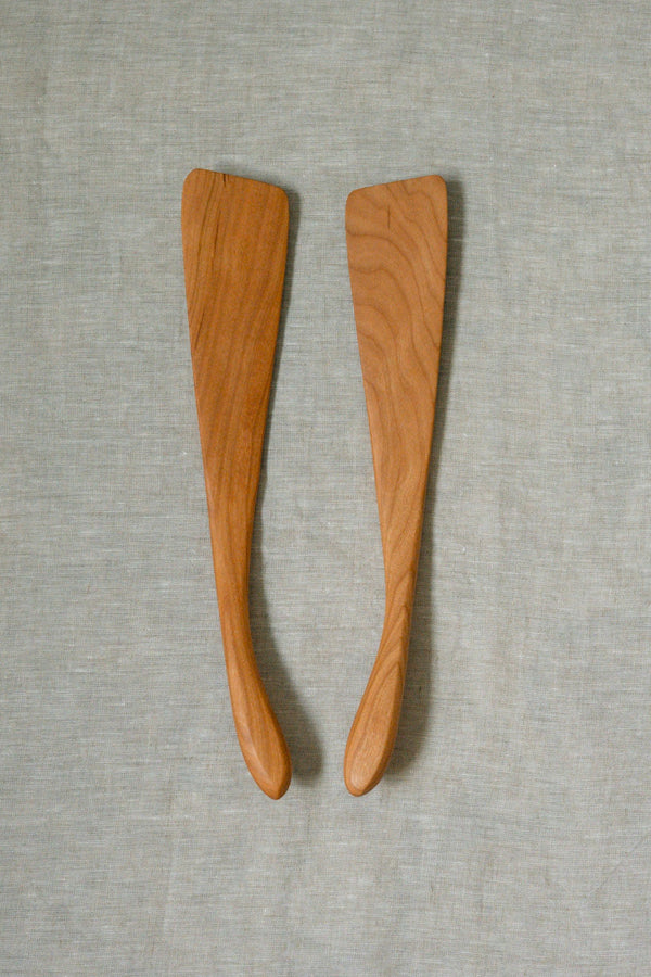 Two cherry wooden french spatulas with visible wood grain, placed side-by-side on a textured light gray fabric background.