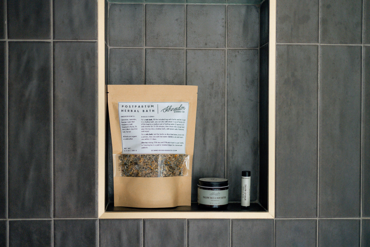 Postpartum herbal sitz bath, calendula tallow balm, and vanilla bean tallow lip balm sit in a black tile shower.