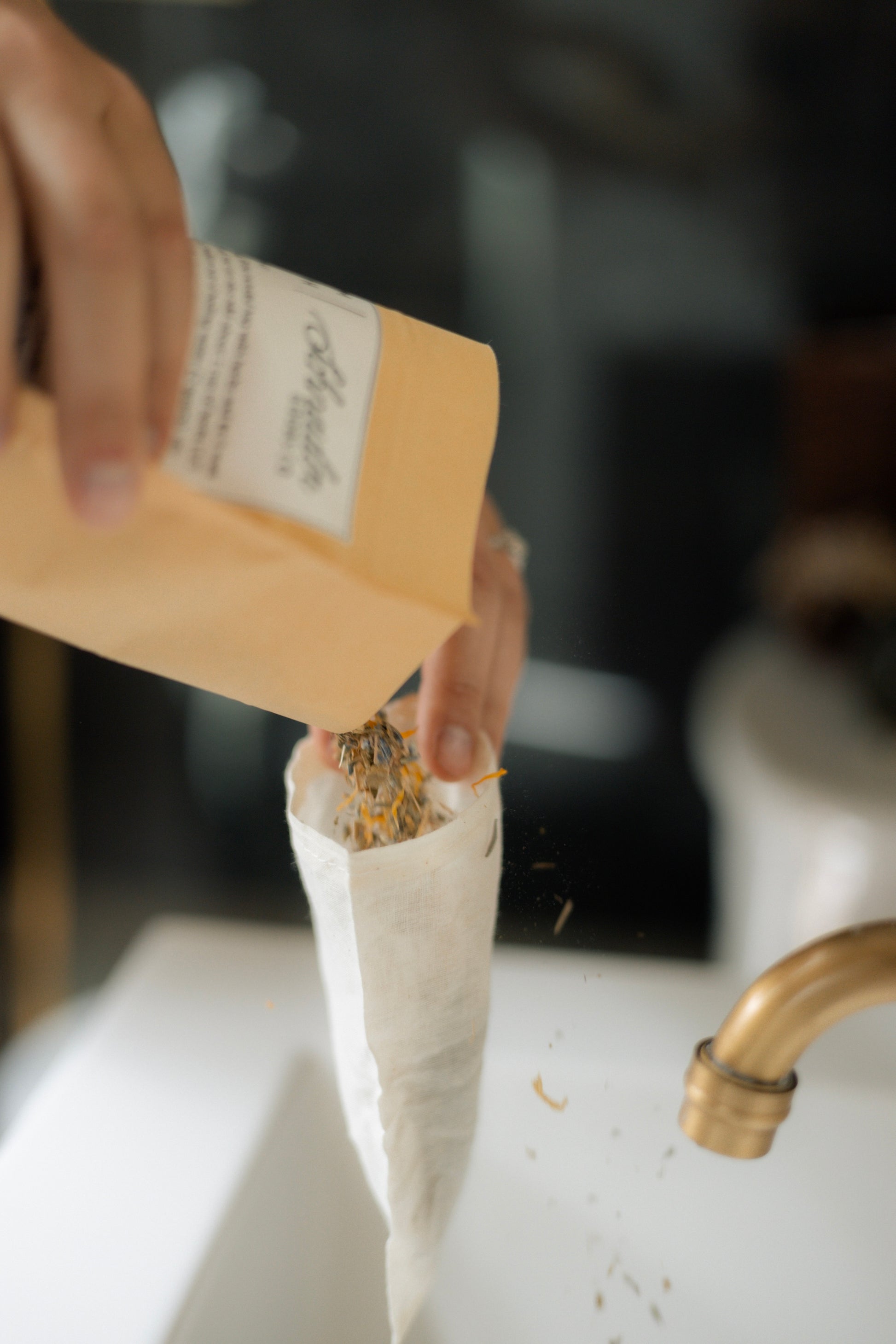 A close-up of a hand pouring dried herbs from a kraft paper pouch into a white muslin bag under a brass faucet.