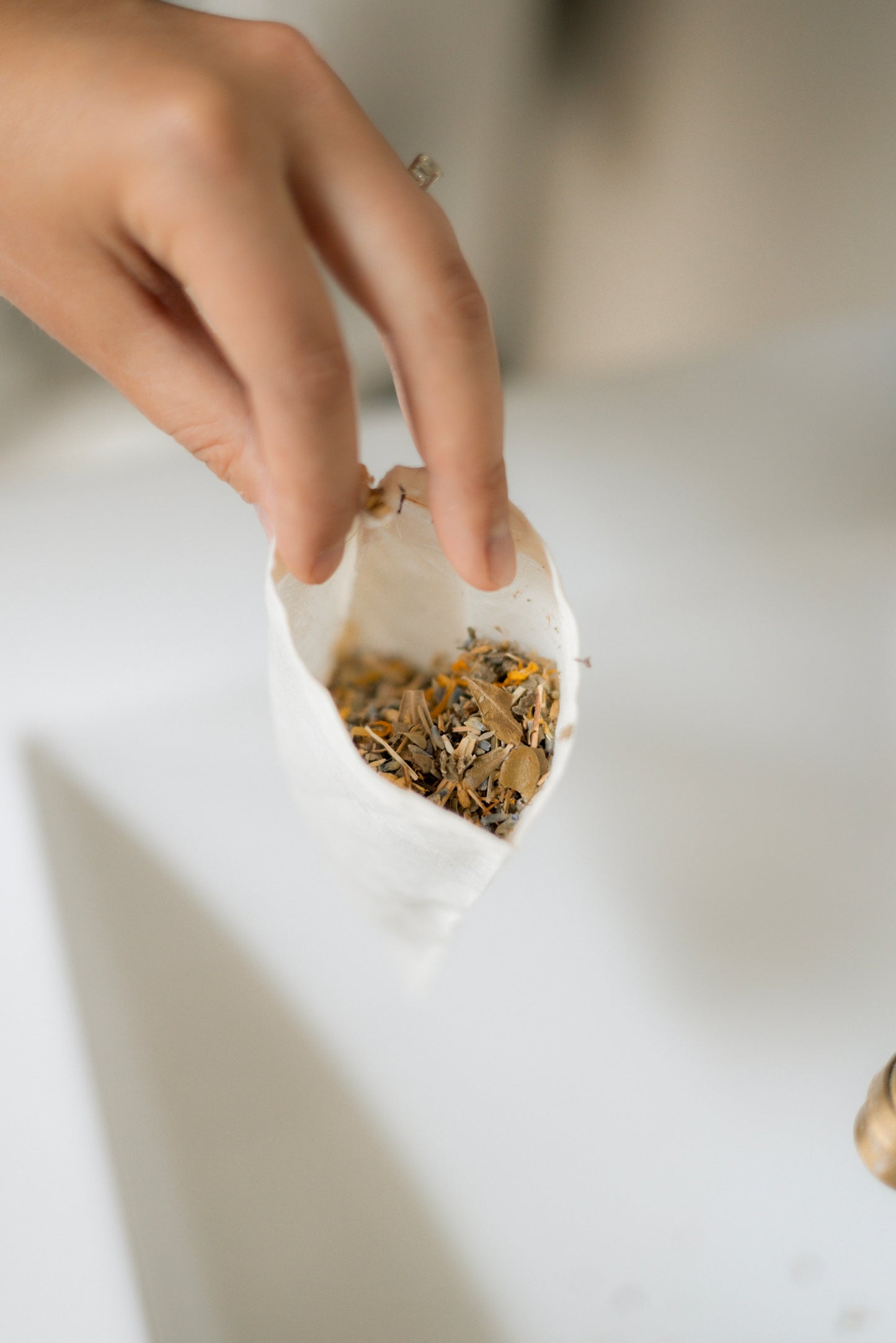 A hand holding the opening of a small white muslin bag filled with dried botanical tea leaves over a white surface.