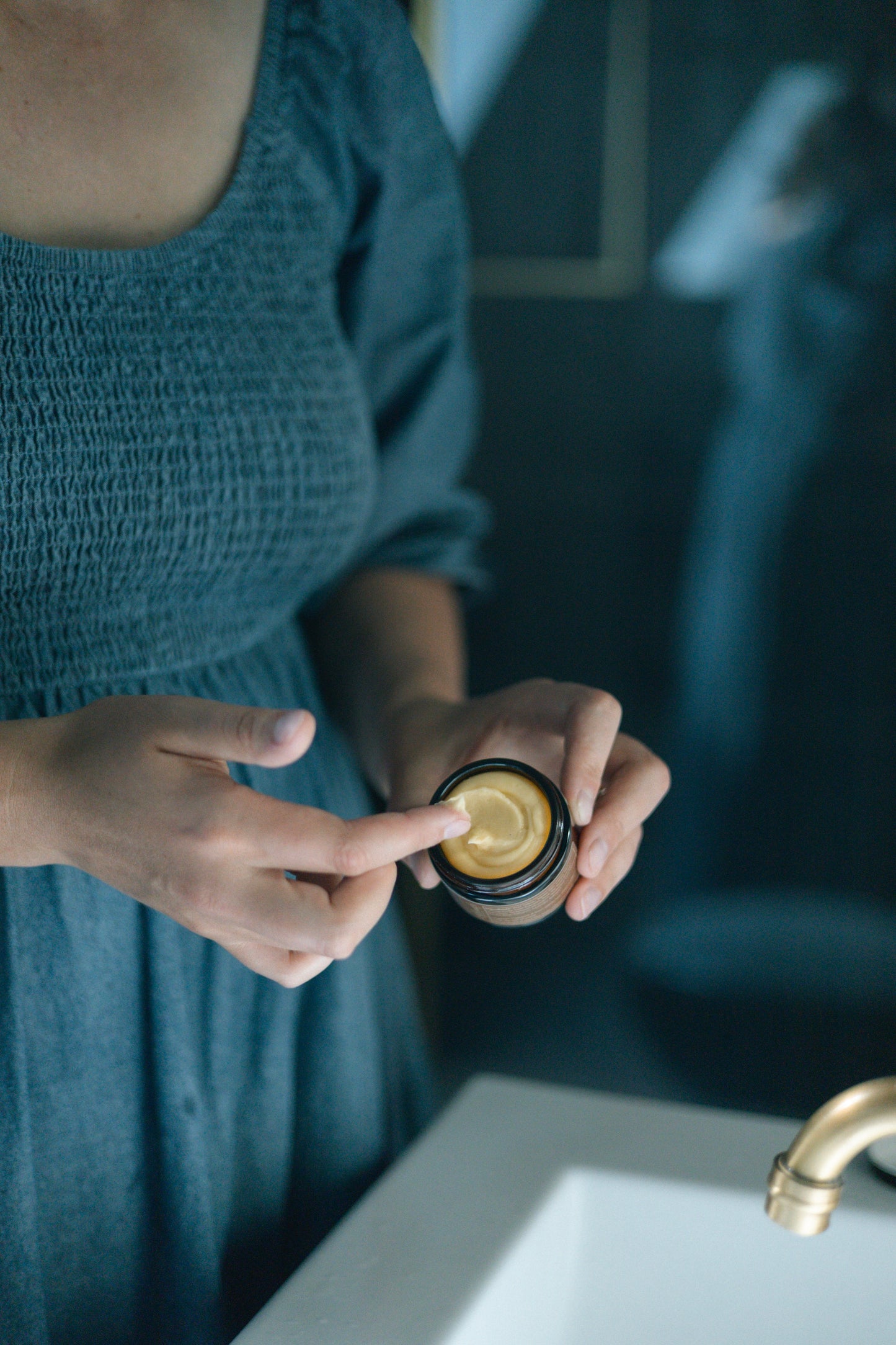 A woman in a blue dress holds Rejuvenate Facial Tallow with Bakuchiol, with a pea-sized amount on her fingertip.