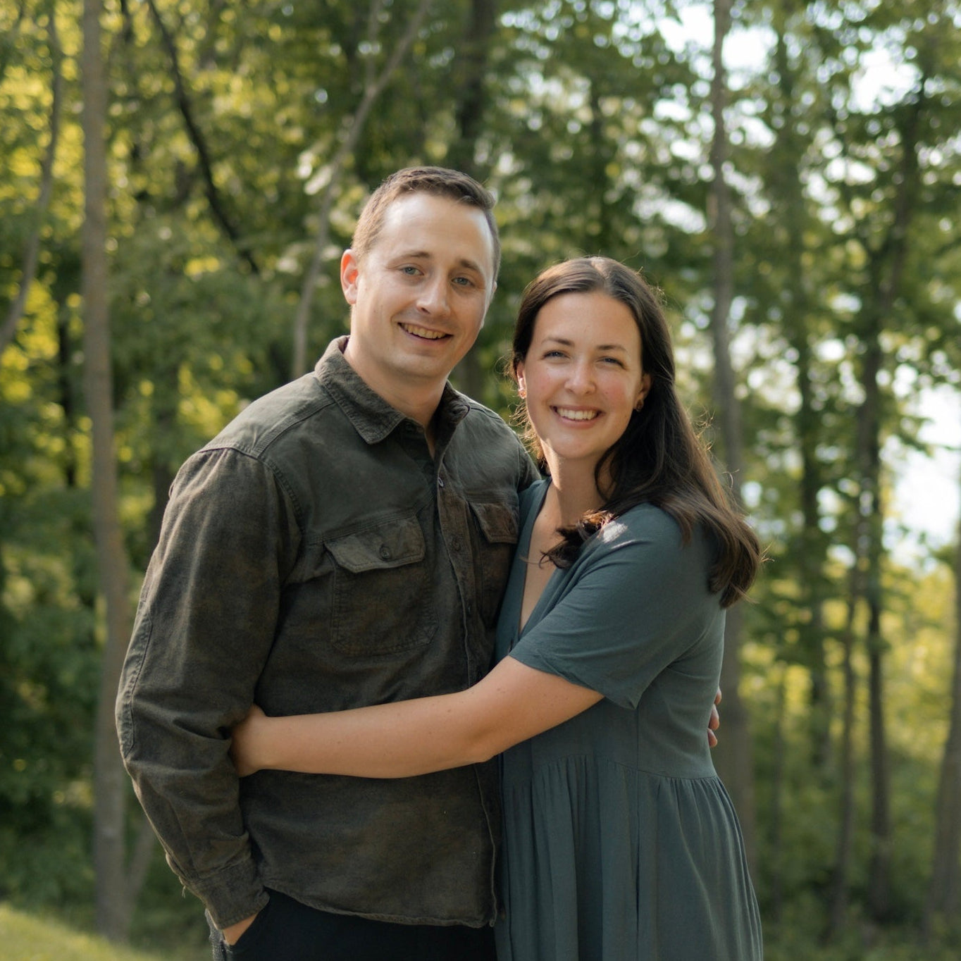 A couple posing together outdoors on a grassy hill with a forest background. The woman is wearing a muted sage green midi dress, and the man is wearing a dark olive green button-up shirt and black pants.