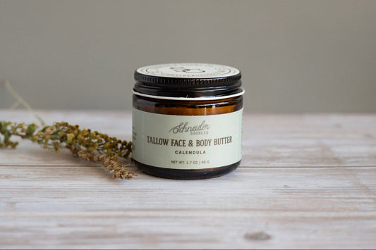 Jar of Schneider Goods Co Calendula Tallow Face & Body Butter on a wooden surface with dried herbs.