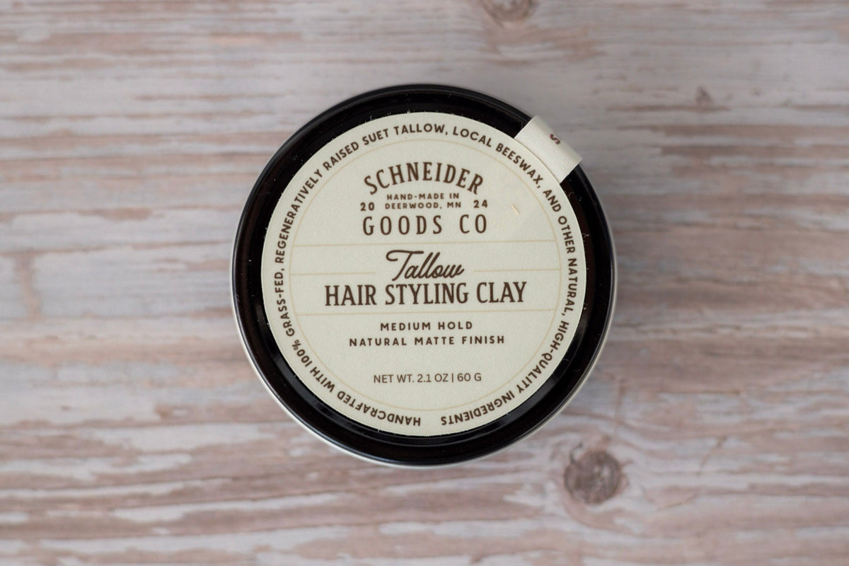 A round tin of Schneider Goods Co. Tallow Hair Styling Clay placed on a light wood background.