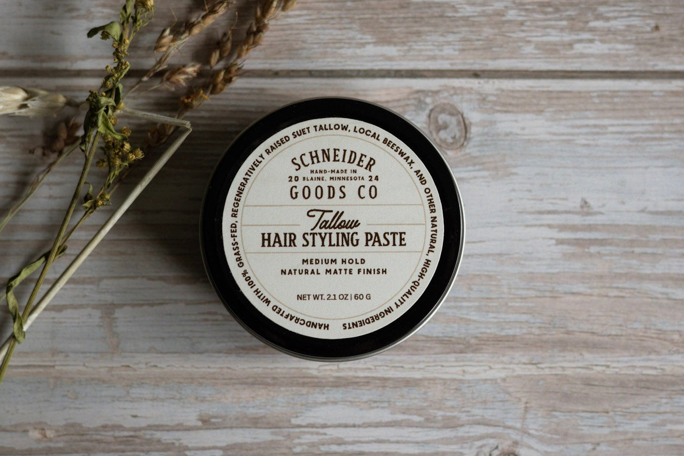 Jar of Schneider Goods Co. hair paste on a wooden surface with dried herbs.