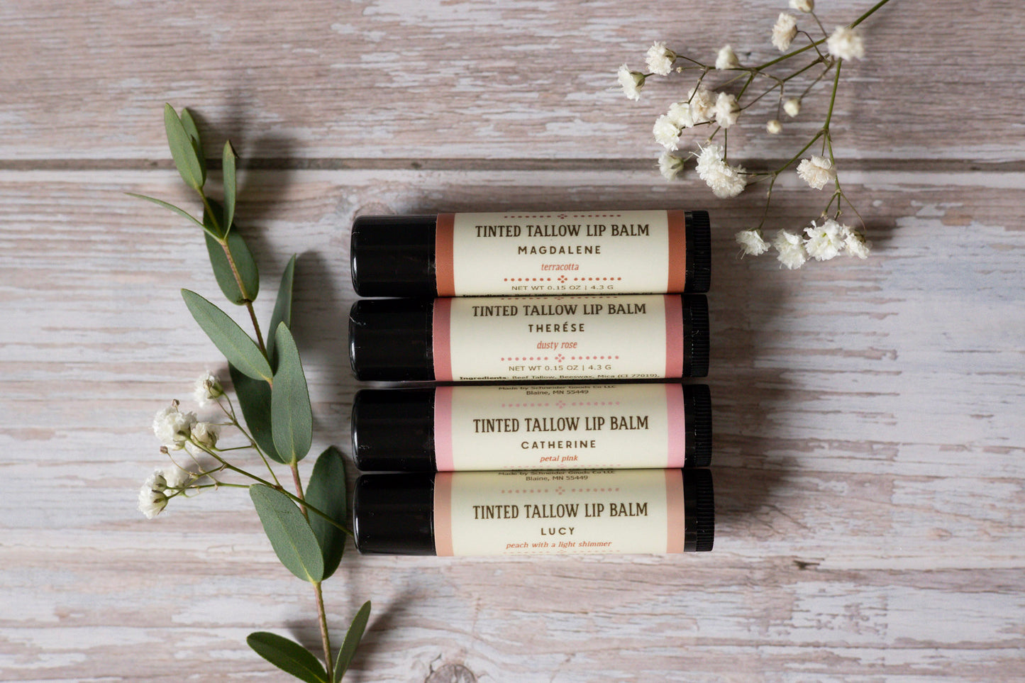 Four tinted tallow lip balms on a wooden surface with flowers and leaves.
