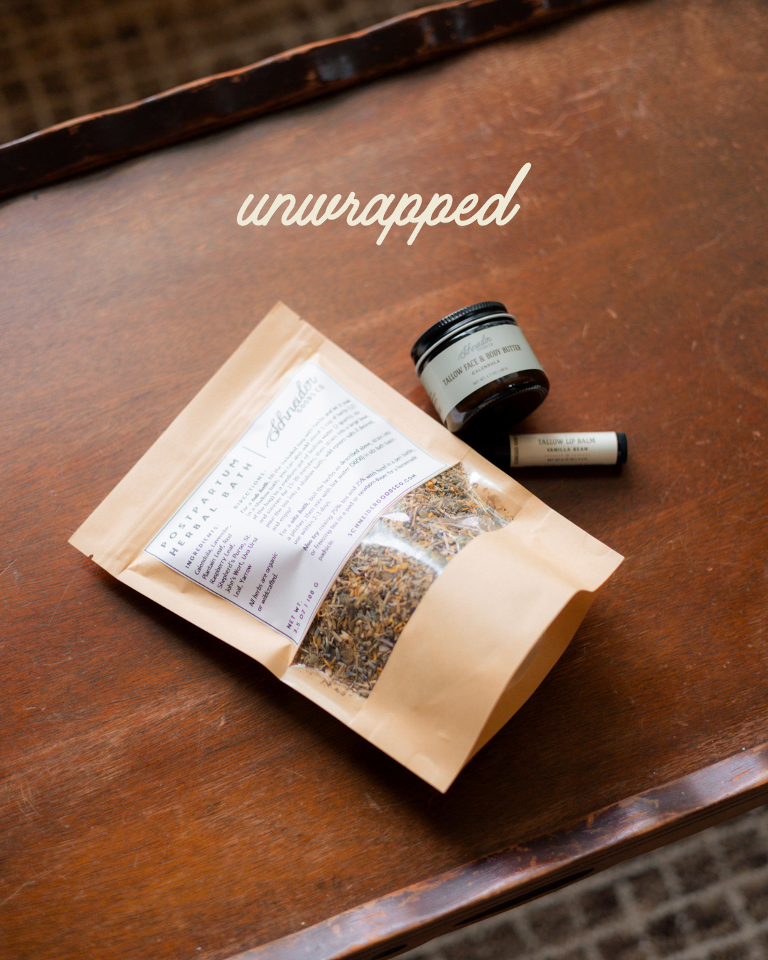 A kraft paper pouch labeled 'unwrapped' with a clear window showing dried botanicals, placed on a dark wooden table next to a small amber jar and a lip balm tube.