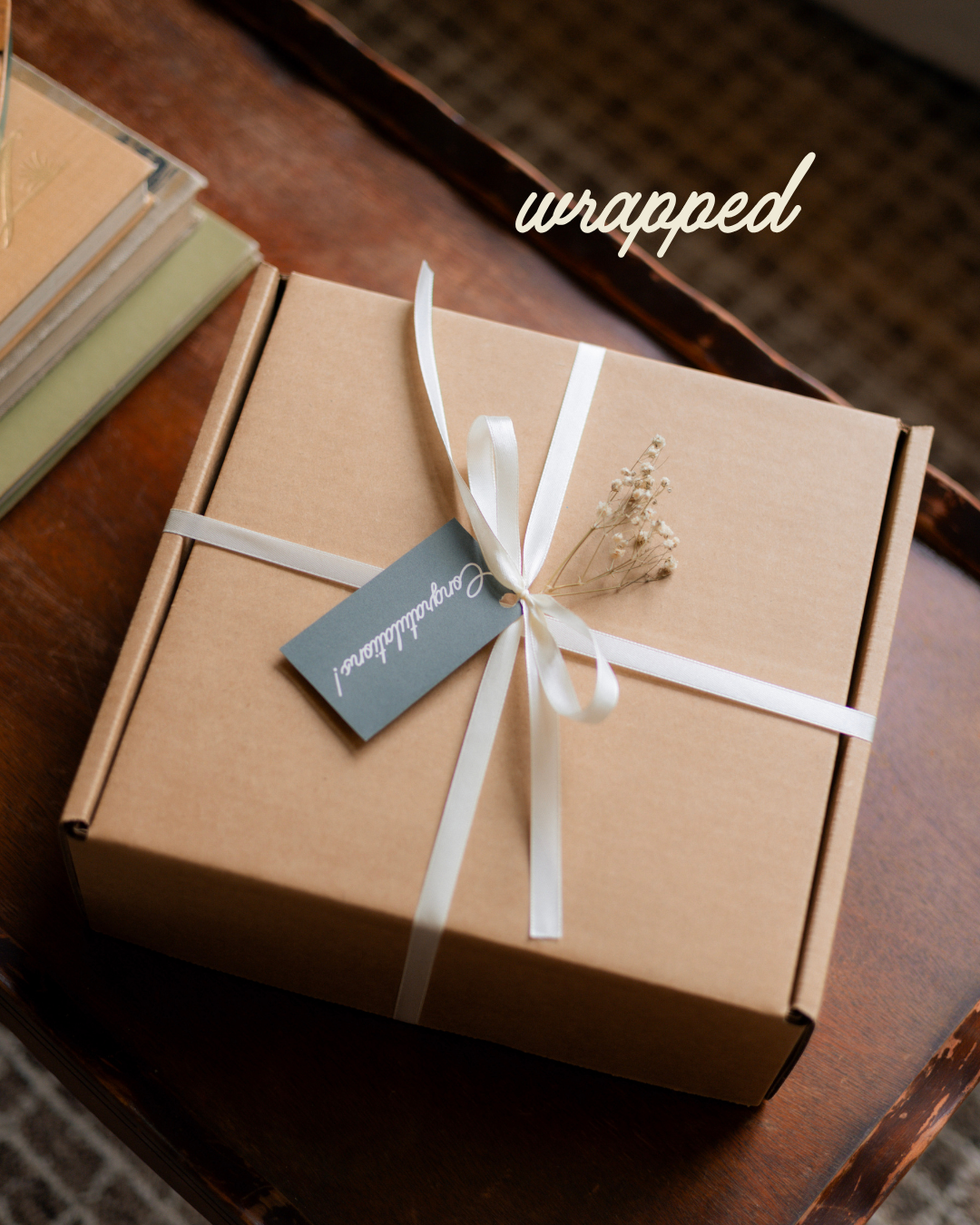 A square kraft paper gift box wrapped with a white ribbon and bow, featuring a small gray tag and dried floral accent, placed on a dark wooden surface with the word 'wrapped' overlaid in the upper right.