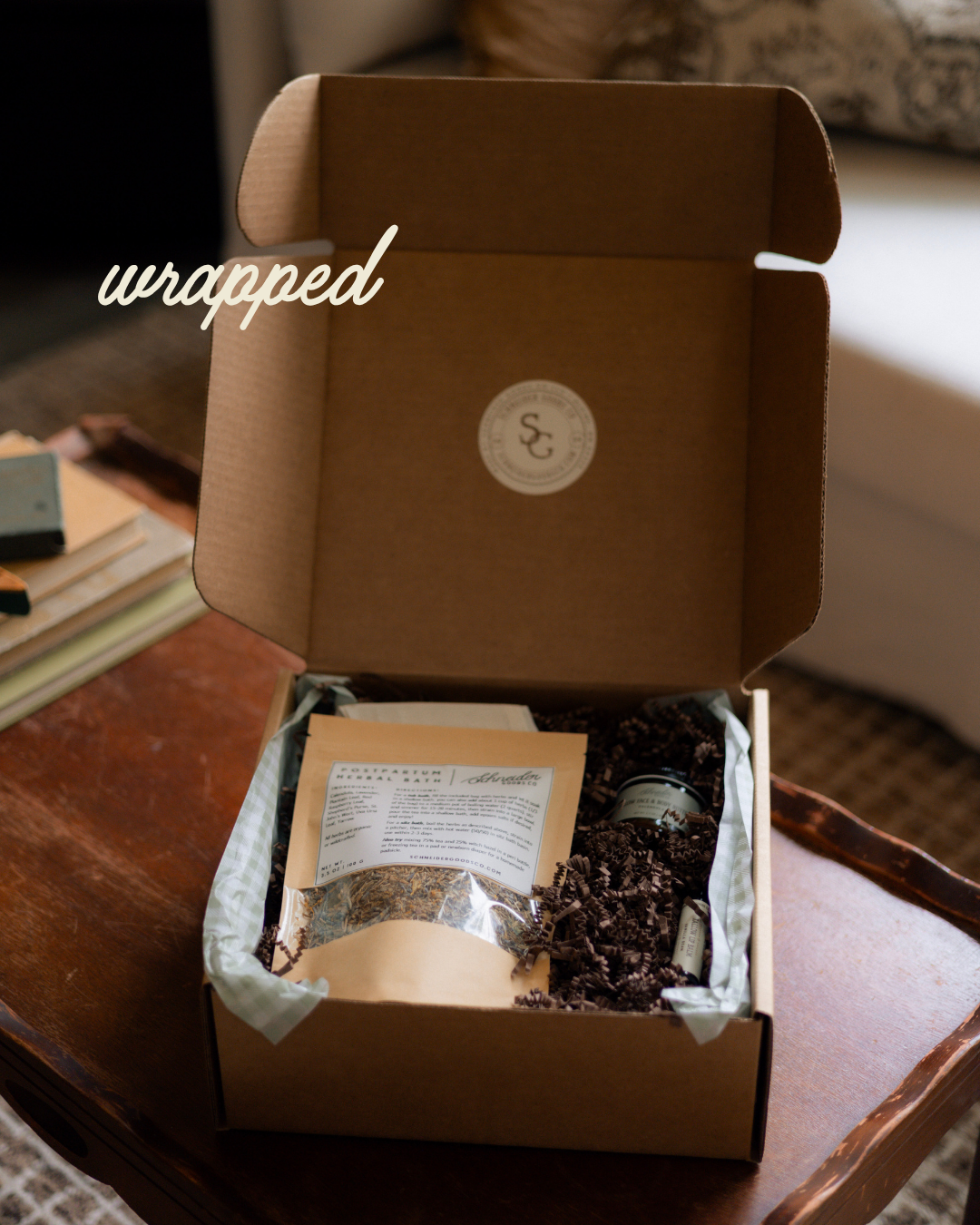 An open brown cardboard gift box sits on a wooden table in a real indoor setting, with a white script overlay reading 'wrapped'. Inside the box are the new mama bundle items, filled with brown krinkle paper, with a circular logo sticker visible on the inside lid.